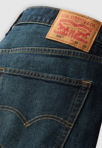 Tmavomodré džínsy z denimu s hnedou koženou záplatou s červeným označením, viditeľným šitím a charakteristickým zakriveným dizajnom zadného vrecka.