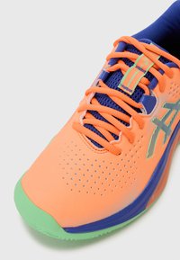 Sportliche Schuhe mit einem orangenen Mesh-Obermaterial und blauen Akzenten, grüner Gummisohle und ausgeprägten Perforationen für Belüftung und Textur.