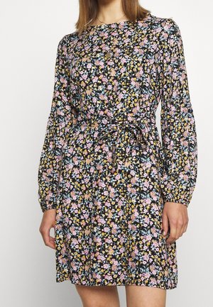 Frau trägt ein langärmeliges schwarzes Kleid mit einem blumigen Muster in Pink, Gelb und Hellblau, an der Taille mit einem Stoffgürtel gebunden.