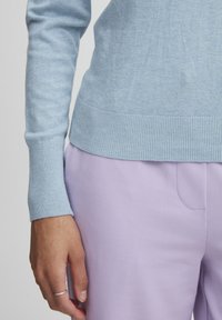 Pull bleu clair avec des poignets côtelés, associé à un pantalon lavande. Le tissu semble doux avec une texture lisse et une légère variation de couleur.