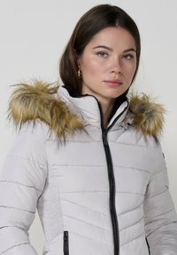 Párnázott fehér dzseki levehető, homokszínű szőrmés kapucnival, cipzáras záródással és oldalsó zsebekkel, fekete részletekkel.