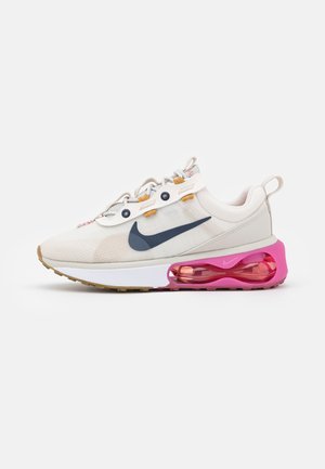 Baskets blanches et beiges avec un swoosh Nike bleu foncé, des passants de lacets orange, une semelle coussinée et une bulle d’air rose bien visible au talon.