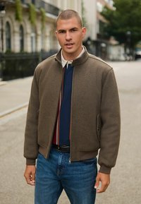 Jachetă bomber cu fermoar de culoare maro, cu mansete ribbate, purtată peste un polo bleumarin cu dungi. Asortată cu blugi albaștri; setting urban în aer liber.