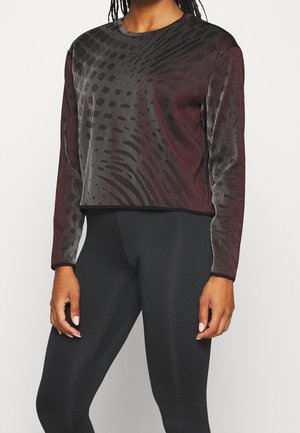 Personne portant un haut à manches longues foncé à motifs et des leggings noirs unis, debout, les bras relâchés le long du corps sur un fond uni.