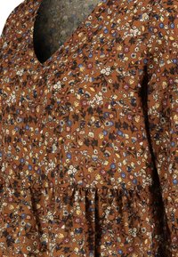 Blumige Bluse mit einem braunen Hintergrund, der mit kleinen weißen, gelben und blauen Blumen geschmückt ist, V-Ausschnitt und gerüschtem unteren Saum.