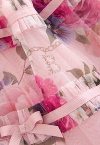 Ziedu mežģīņu audums rozā krāsā, ar dzīvīgām ziedu apdrukām un smalku ķēdes rakstu. Akcentēts ar gaiši rozā lentu, kas sasieta bantē.