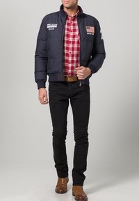 Navy puffer jacket with American flag patch, gray accents, worn over a red plaid shirt, paired with black jeans and brown boots.