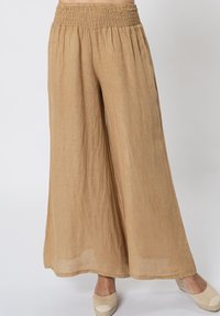 Pantaloni in lino beige a gamba larga con una vita arricciata e una finitura morbida e strutturata. Accentuati da calzature leggere e casual.