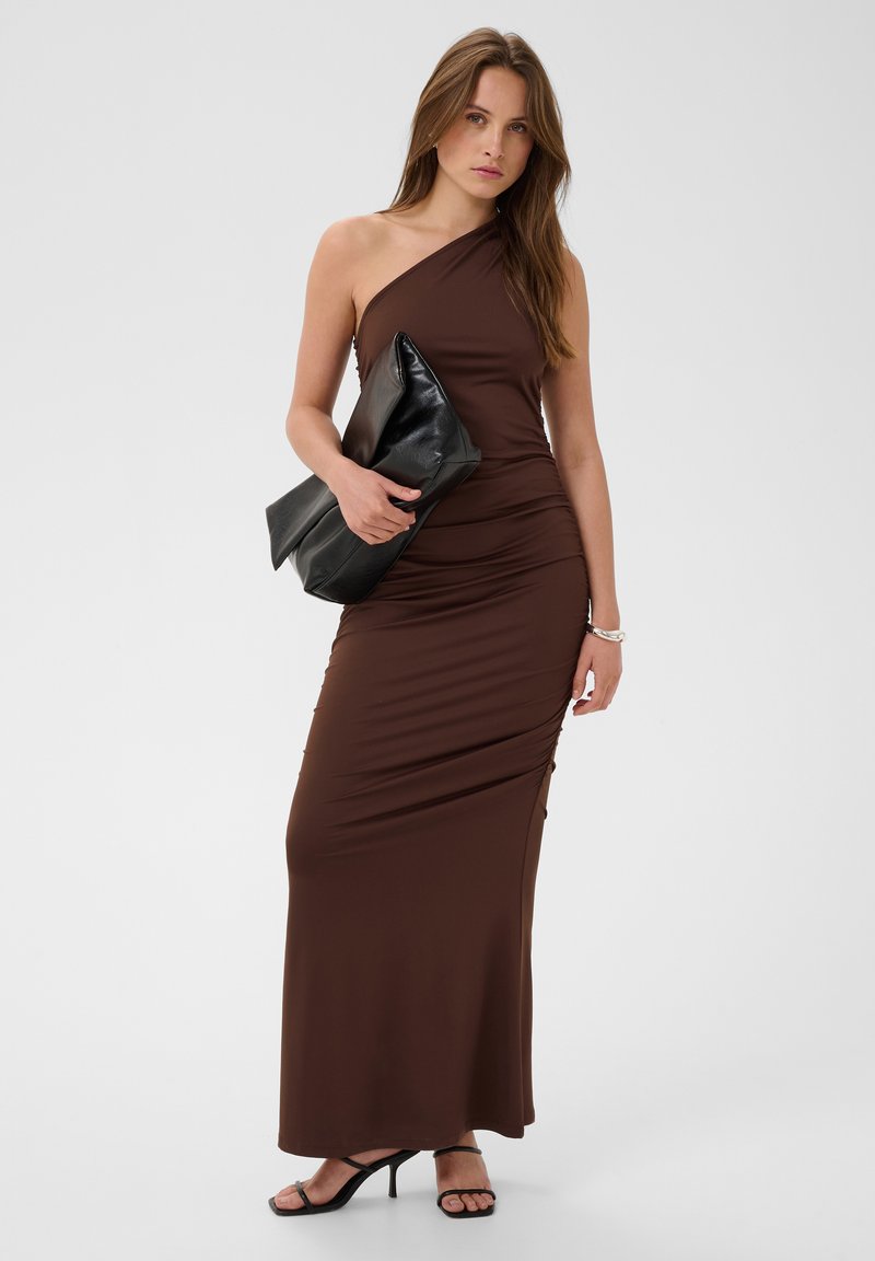 Femme portant une robe maxi marron à une épaule, tenant une grande pochette noire, et portant des sandales à talons noires sur un fond uni.