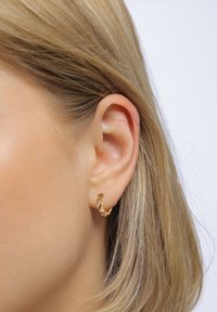 Goldfarbene Creolen mit einem gedrehten Design, das drei kleine klare Edelsteine zeigt. An einem Ohr positioniert, nahe glattem, blondem Haar.