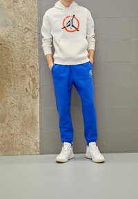 Felpa con cappuccio da uomo grigio chiaro con logo, abbinata a pantaloni della tuta blu brillante. Scarpe da ginnastica bianche completano il look. Design semplice, vestibilità casual.