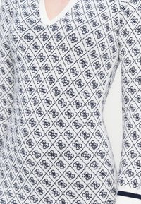 Tuvredzēts balts adīts džemperis ar tumši zilām dimantu rakstām un savstarpēji savienotām monogrammām, V veida kakla izgriezums, kā arī tumši zilas svītras uz piedurknēm.