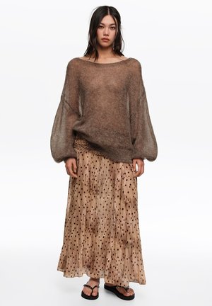 Kobieta ubrana w luźny brązowy przezroczysty sweter, długą beżową spódnicę w ciemne grochy oraz czarne sandały na platformie, stoi na białym tle.