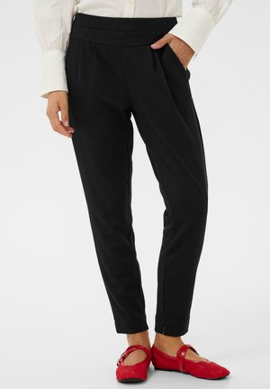 Sorte skræddersyede bukser med plisseringer, lavet af et struktureret stof. Kombineret med røde flade sko med en rem og sløjfedetalje.