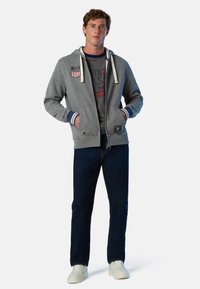 Uomo in piedi con le mani in tasca, indossando una felpa grigia con zip e patch degli Stati Uniti, jeans scuri, sneakers bianche e una t-shirt stampata sotto.