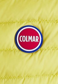 Röd cirkulär gummilogotyp med vit text "COLMAR" utritad i blått på en gul quiltad tygbakgrund.