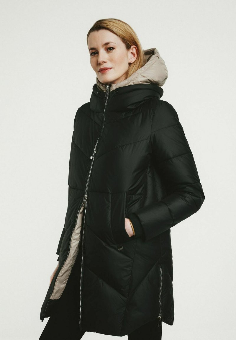 Schwarze Pufferjacke mit geometrischem Steppmuster und grauem Kapuzenansatz. Sie verfügt über einen Frontreißverschluss und seitliche Taschen. Aus warmem und isoliertem Material.
