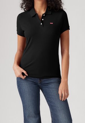 Vrouw draagt een zwart poloshirt met korte mouwen, witte knopen en een klein rood Levi's-logo, gecombineerd met een blauwe spijkerbroek.