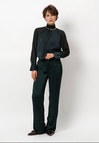 Zwarte zijden blouse met een hoge hals en gouden accent, gecombineerd met donkergroene broek met wijde pijpen en een gestructureerde afwerking.