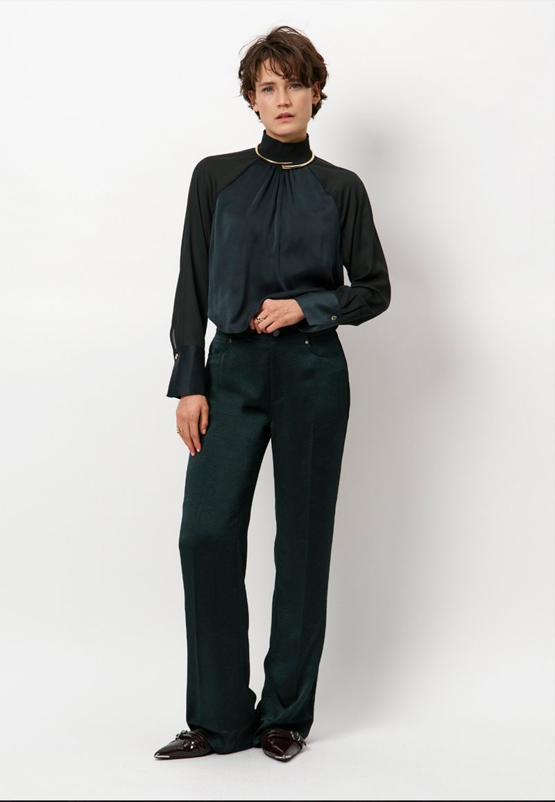 Schwarze Seidenbluse mit hohem Kragen und goldenen Akzenten, kombiniert mit dunkelgrünen weit geschnittenen Hosen mit strukturiertem Finish.