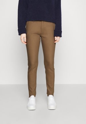 Person iført tætsiddende brune bukser, navyblå sweater og hvide sneakers, der står foran en ensfarvet lys baggrund.