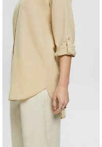 Beige, lightweight shirt with rolled sleeves and a slightly curved hem, paired with light-colored, loose-fitting pants.