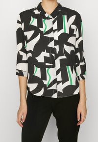 Blouse noir et blanc à motifs géométriques, avec des accents verts. Tissu léger, fermeture à boutons et manches trois-quarts.