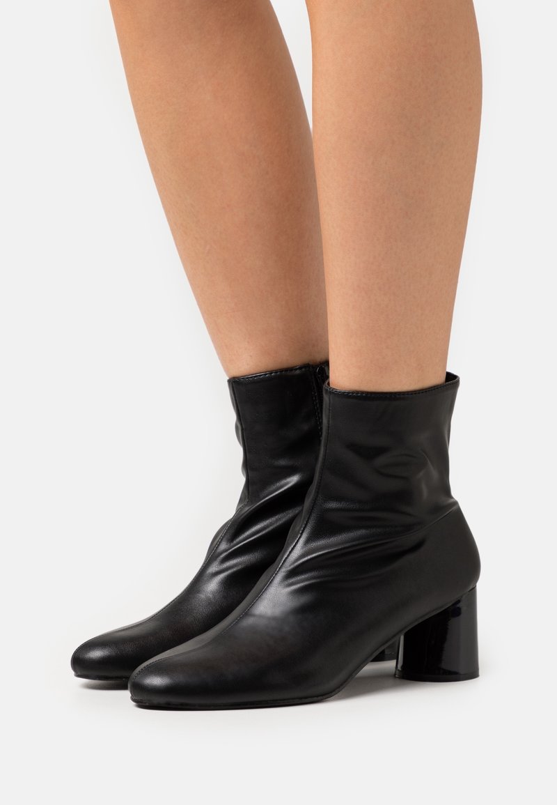 Bottes en cuir noir montant à la cheville avec talons carrés, portées sur des jambes nues, vues de côté sur fond blanc.