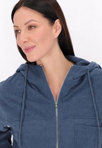 Marineblauwe gewatteerde jas met capuchon, ritssluiting, voorzak en trekkoord. Beschikt over verticale strepen en metalen ritsaccenten.