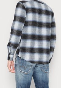 Hombre vistiendo una camisa de cuadros en negro, blanco y azul de manga larga combinada con jeans azules desgastados, mostrado desde la espalda y a partir de la cintura.