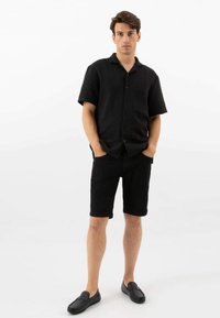 Zwarte korte mouwen overhemd met een textuur, gecombineerd met zwarte shorts. Zwarte instappers maken de outfit compleet. Effen witte achtergrond.