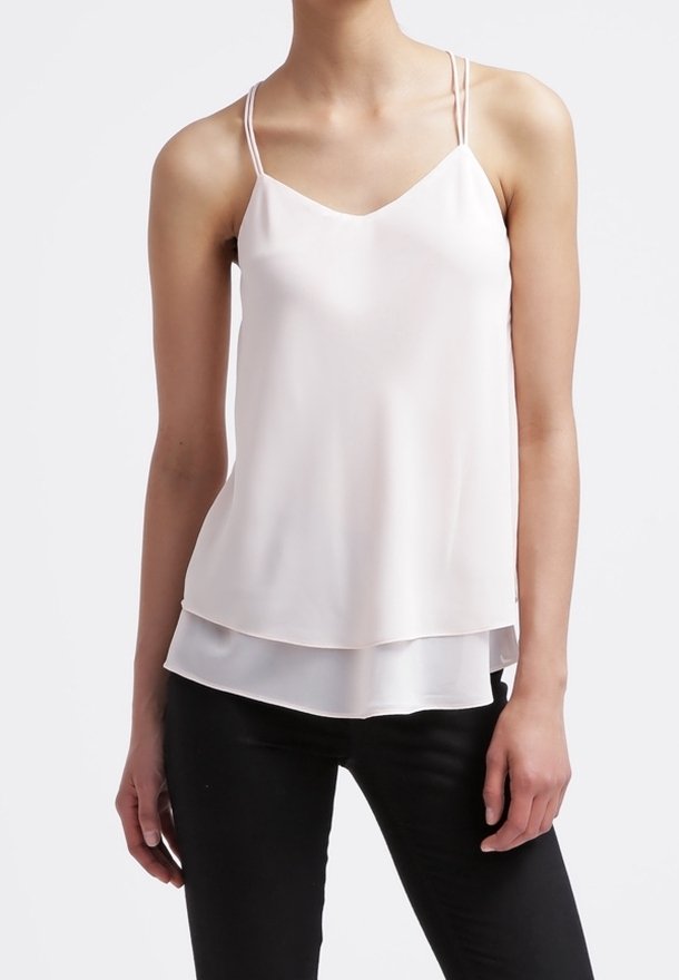 Kobieta w bezrękawnikowej, białej, warstwowej bluzce na cienkich podwójnych ramiączkach oraz czarnych spodniach, stojąca na tle jednorodnego białego tła.