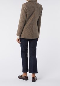 Personne portant une veste marron texturée, un jean droit bleu foncé et des mocassins marron foncé, debout devant un fond clair uni.