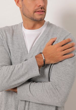 Grå cardigan lavet af blødt stof, båret over en hvid t-shirt. Sort læderarmbånd med en sølvaccent på håndleddet.