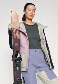 Skijacke in Hellgrau und Pink, graues Oberteil und lila Skihosen; hält einen dunklen Ski mit weißen Bindungen und Logodetails.