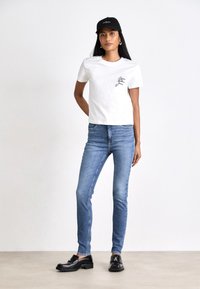 Fehér pamut póló fekete logóval, világoskék farmer skinny nadrág, fekete mokaszin, és fekete sapka. Egyszerű háttér.
