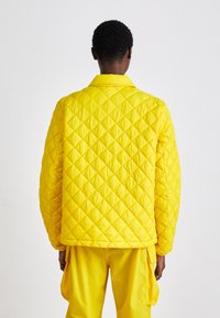 Casaco amarelo acolchoado com um padrão de diamante, com gola e um corte descontraído. O material parece liso e almofadado para isolamento.