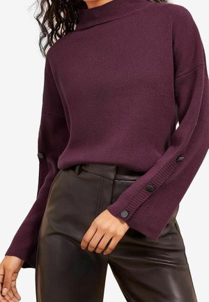Lichtgewicht, geribbelde trui in diep paars met een mock-halslijn en uitlopende mouwen, met zwarte knoopdetails aan de manchetten. Gecombineerd met zwarte broek.