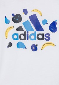 Maglietta bianca in cotone con una grafica colorata di frutta e il logo "adidas" in blu e nero, con design giocosi e facce sorridenti.
