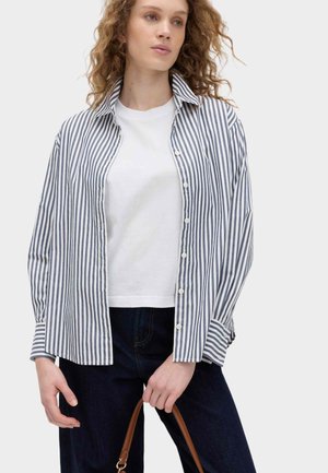 Vrouw met krullend haar, gekleed in een blauw-wit gestreept overhemd met knopen over een wit T-shirt, donkere spijkerbroek, die een bruine handtasriem vasthoudt.