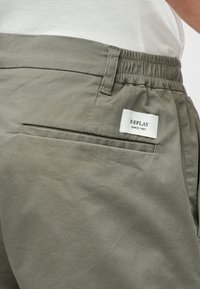 Gros plan sur un pantalon vert olive avec une taille élastique, une poche arrière, une passante de ceinture et une étiquette blanche indiquant "REPLAY SINCE 1981" sur le côté.