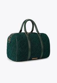 Pikowana zielona torba materiałowa z skórzanymi uchwytami i akcentami, posiadająca zamek błyskawiczny i prostokątny kształt. Widoczne detale w kolorze złotym.