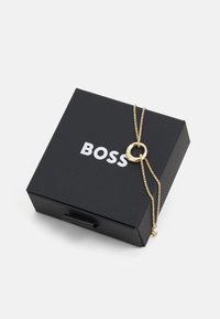 Collana d'oro con pendente circolare, esposta su una scatola nera con logo "BOSS" in bianco. La catena presenta una texture fine e piccoli dettagli.