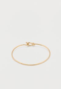 Λεπτό γυαλιστερό χρυσό βραχιόλι με καμπύλο βρόχο σύρματος και κούμπωμα με στρίψιμο σε απλό ανοιχτό φόντο.