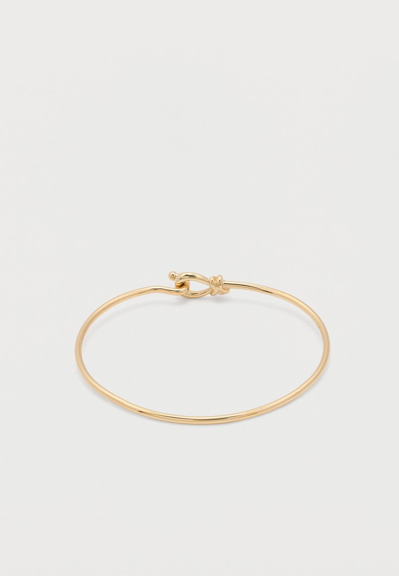 Λεπτό γυαλιστερό χρυσό βραχιόλι με καμπύλο βρόχο σύρματος και κούμπωμα με στρίψιμο σε απλό ανοιχτό φόντο.