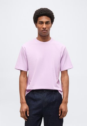 Άνδρας με κοντά κατσαρά μαλλιά που φοράει ένα ανοιχτό μοβ κοντομάνικο μπλουζάκι και σκούρο παντελόνι, στέκεται μπροστά σε ένα απλό φωτεινό φόντο.