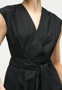 Γυναίκα που φοράει μαύρο αμάνικο φόρεμα τύπου wrap με βαθύ V ντεκολτέ και ζώνη στη μέση, εμφανίζεται από το λαιμό έως τους γοφούς σε λευκό φόντο.