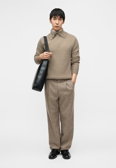 Μπεζ πλεκτό πουλόβερ με ψηλό γιακά, καφέ πλισέ παντελόνια και μαύρα δερμάτινα παπούτσια. Κρατάει μια μαύρη τσάντα ώμου. Καθαρό λευκό φόντο.