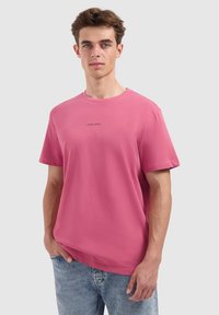 Rosa Baumwoll-T-Shirt mit Rundhalsausschnitt und kurzen Ärmeln, auf der Vorderseite mit dem Text "PURE PATH" bedruckt. Kombiniert mit hellblauen Jeans.