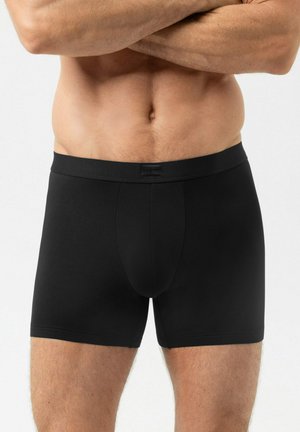 Männlicher Oberkörper mit verschränkten Armen, bekleidet mit schlichten schwarzen Boxershorts, vor neutralem Hintergrund.